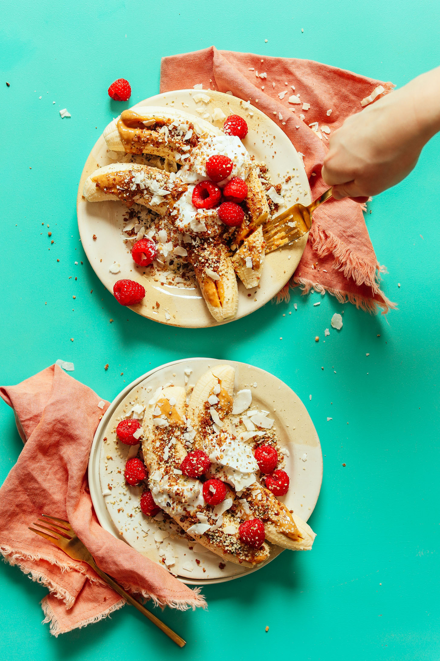 Two serving plates of Raw Banana Splits topped with peanut butter, coconut yogurt, raspberries, flaked coconut, and rawnola
