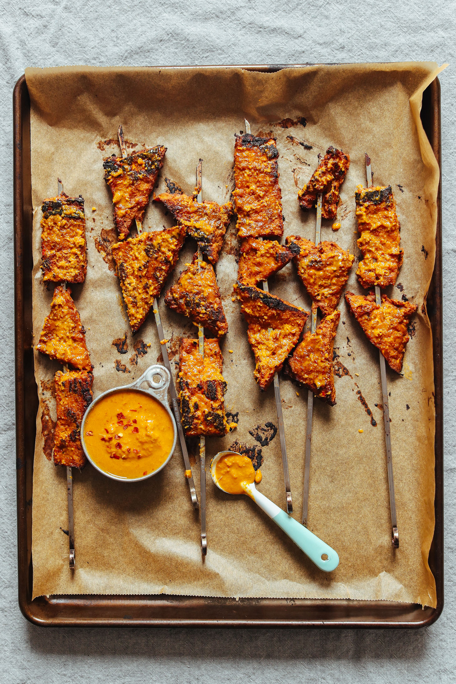 Parchment-lined baking sheet with gluten-free vegan Lemongrass Tempeh Satay skewers and dipping sauce