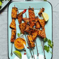 Tray of Vegan Tempeh Satay skewers alongside lime wedges, cilantro, and peanut sauce