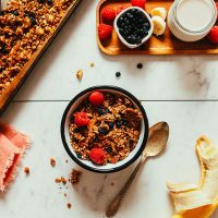 Bowl of vegan Buckwheat Granola alongside fresh fruit, dairy-free milk, and a baking sheet with more granola