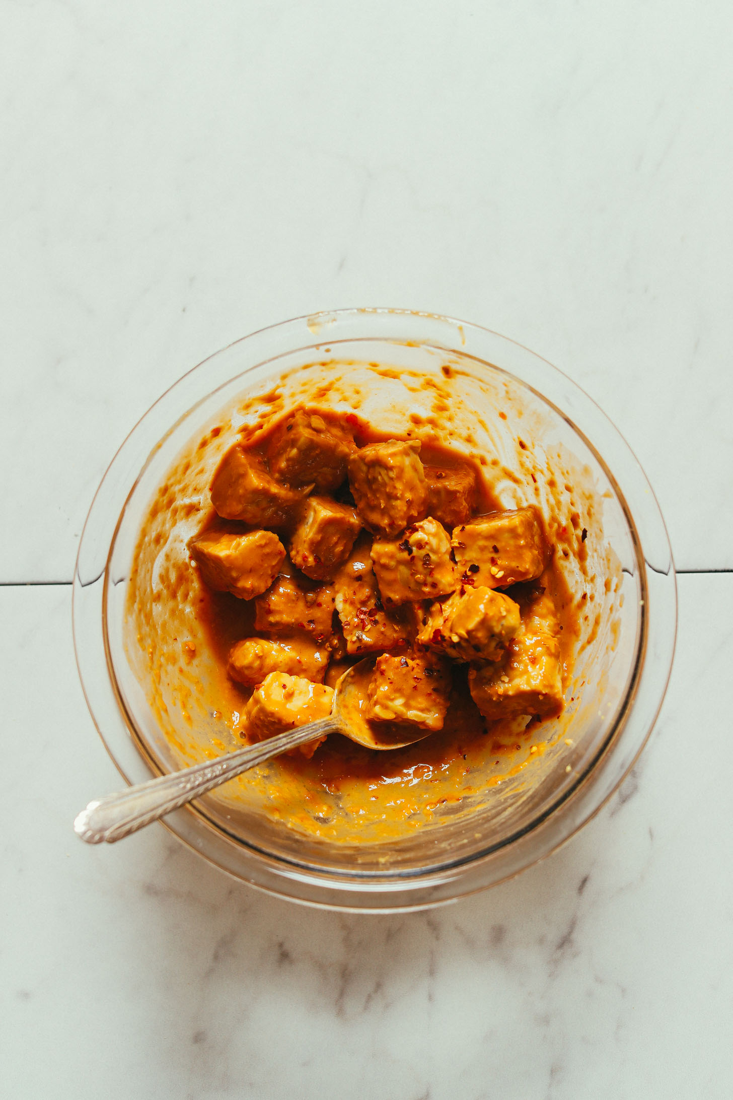 Overhead image of bowl with tempeh marinating in peanut sauce and a spoon for stirring