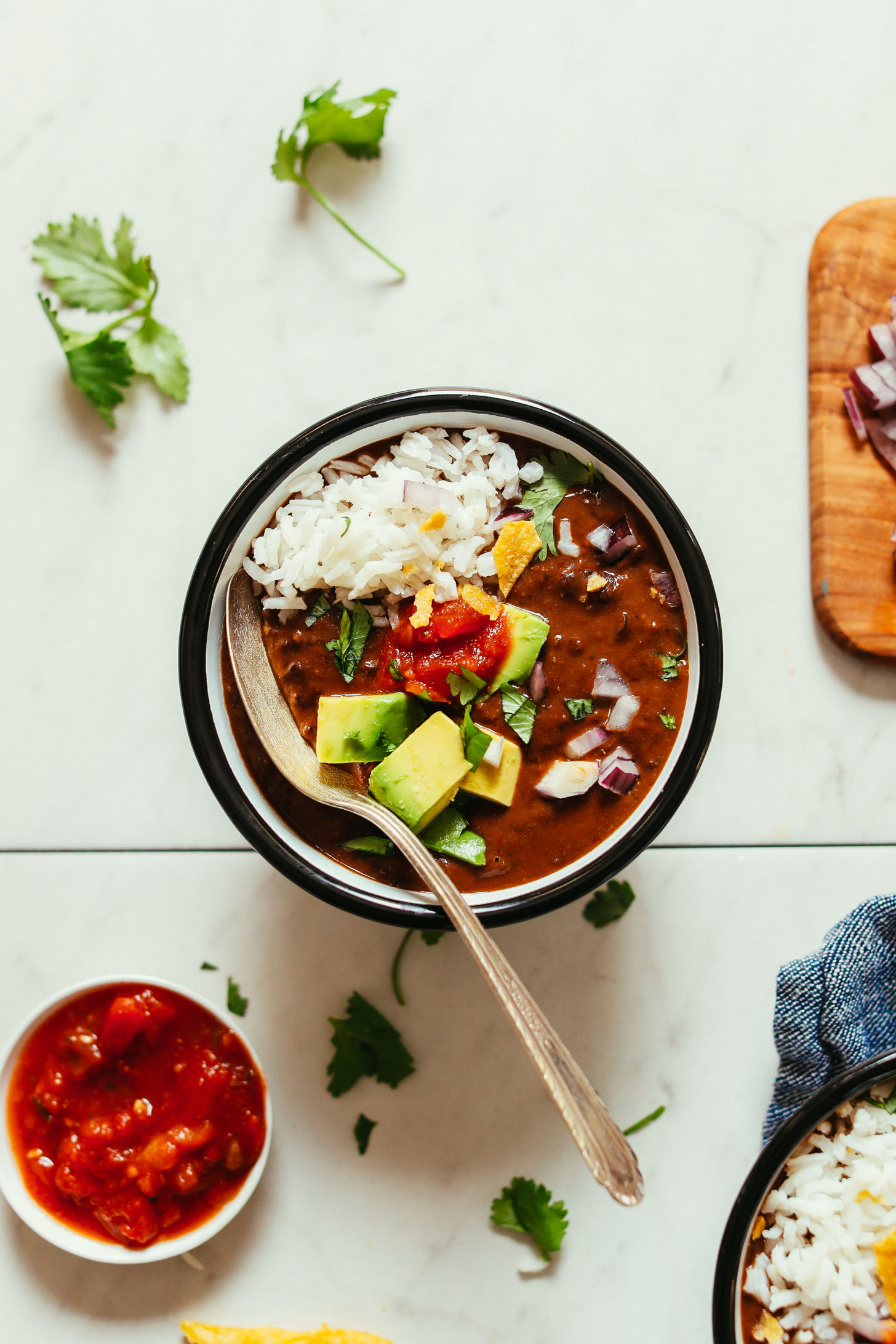Overhead image of black bean soup in a black bowl with a spoon resting in the bowl, garnished with rice avocado, salsa, and chips
