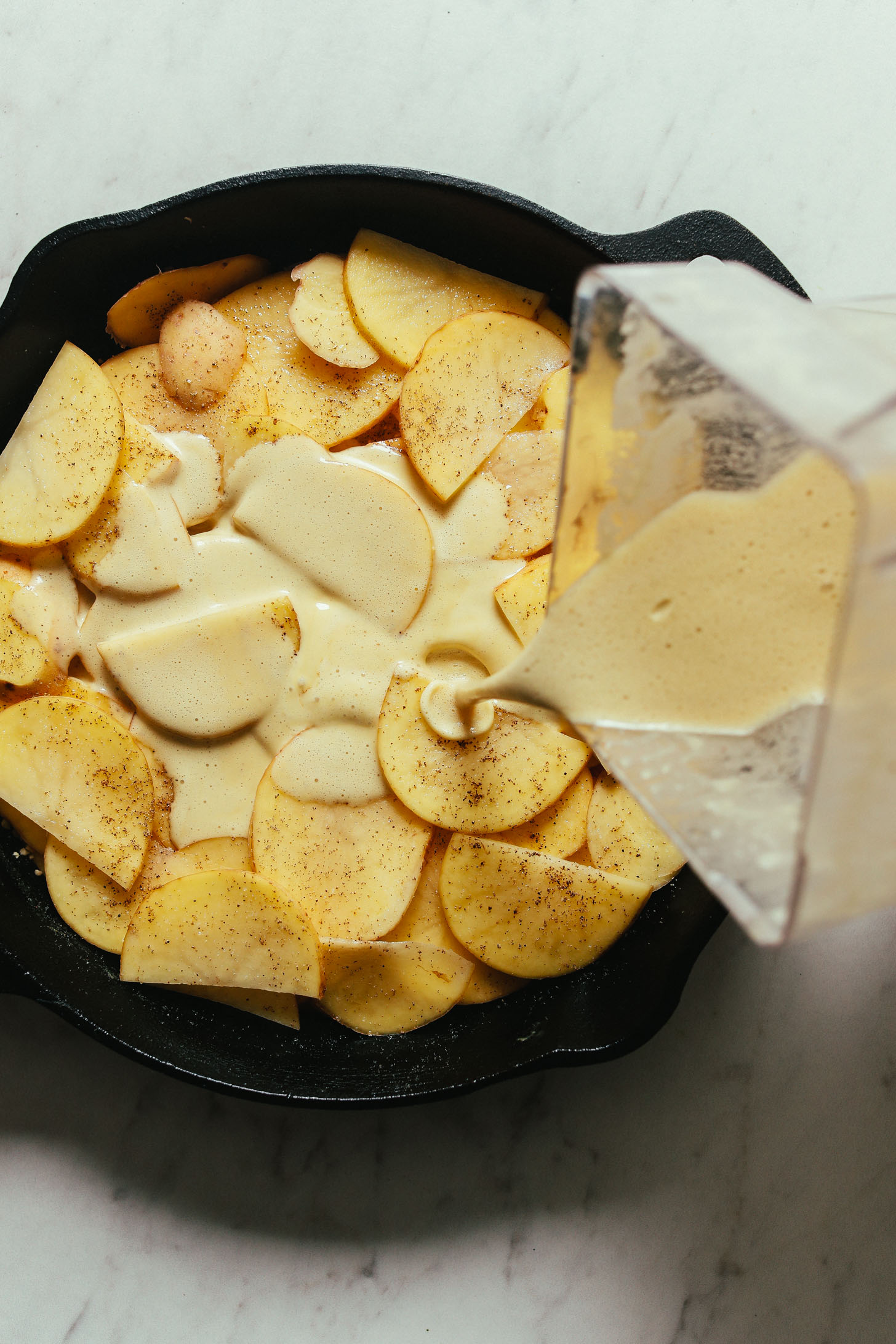 Pouring creamy sauce into a cast-iron skillet for Vegan Scalloped Potatoes