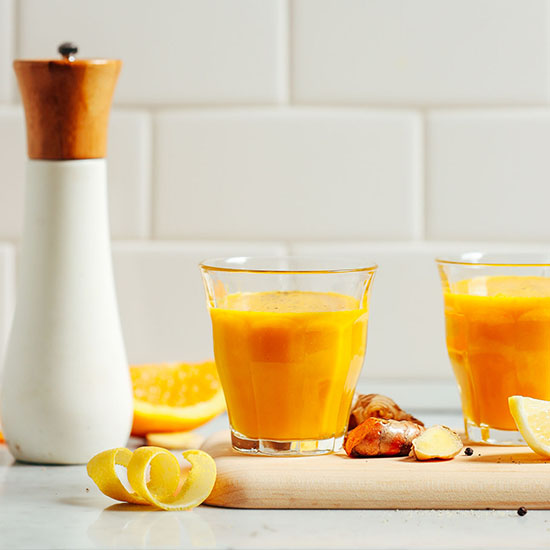 Glasses of our Lemon Ginger Turmeric Wellness Shots on a cutting board with ingredients to make them