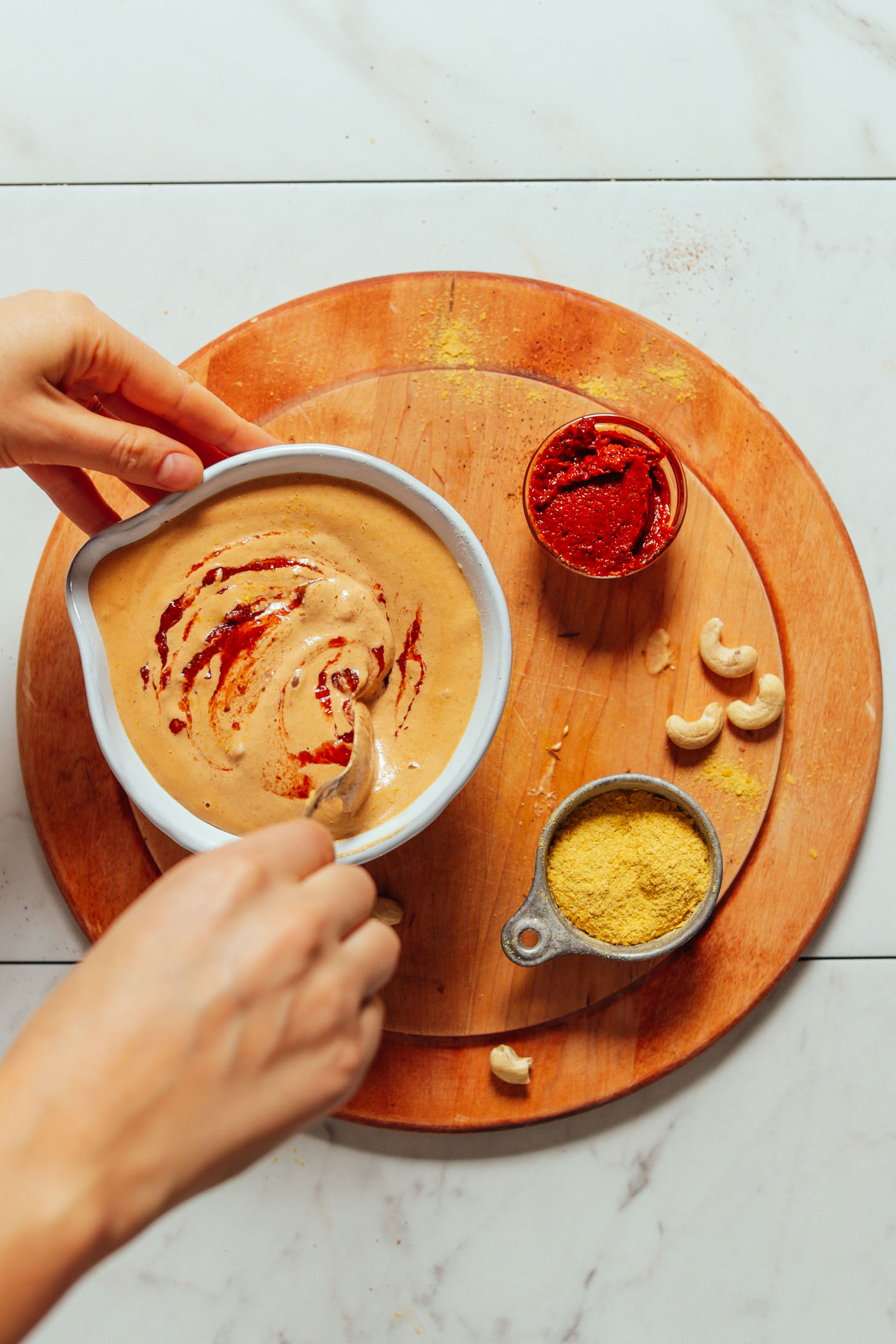 Overhead image of vegan cashew queso in a white bowl with a hand stirring some hot sauce on top and cashews, harissa, and nutritional yeast on the side