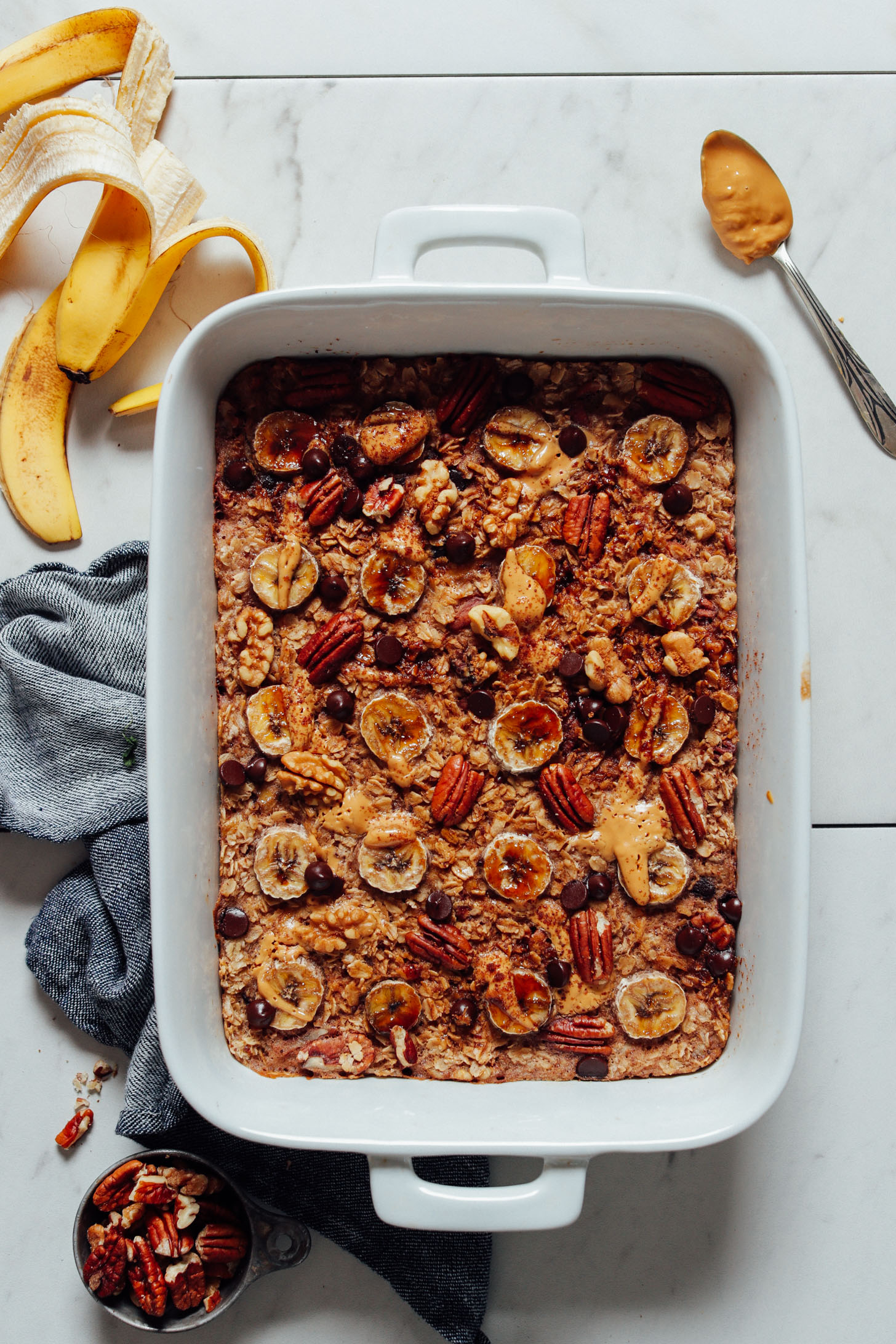 Overhead image of banana baked oatmeal with nuts and dark chocolate with nut butter and banana on the side