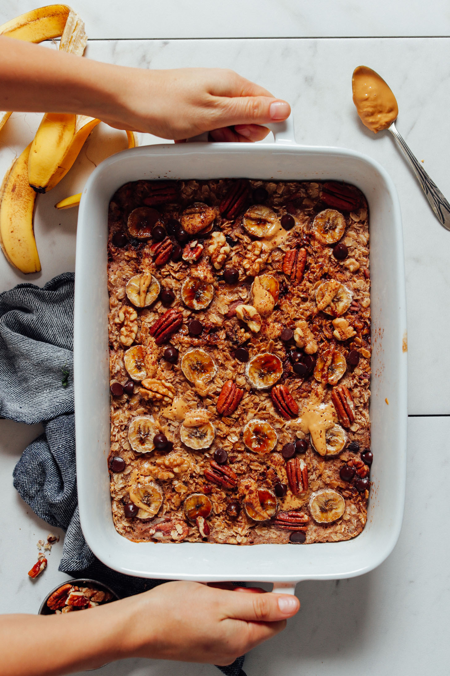 Overhead image of banana baked oatmeal in baking dish with hands holding the pan, and banana and nut butter on the side