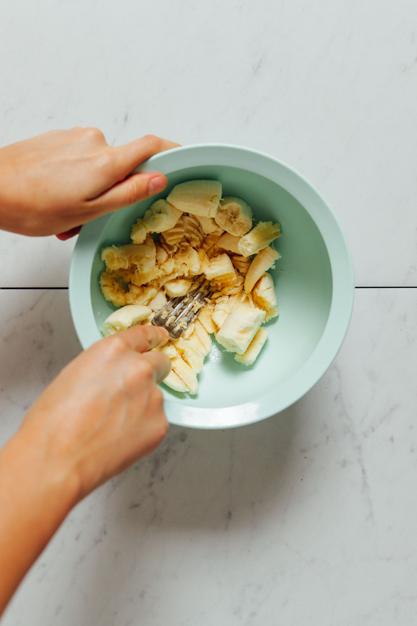 Overhead image of bananas being smashed with a fork in a teal bowl