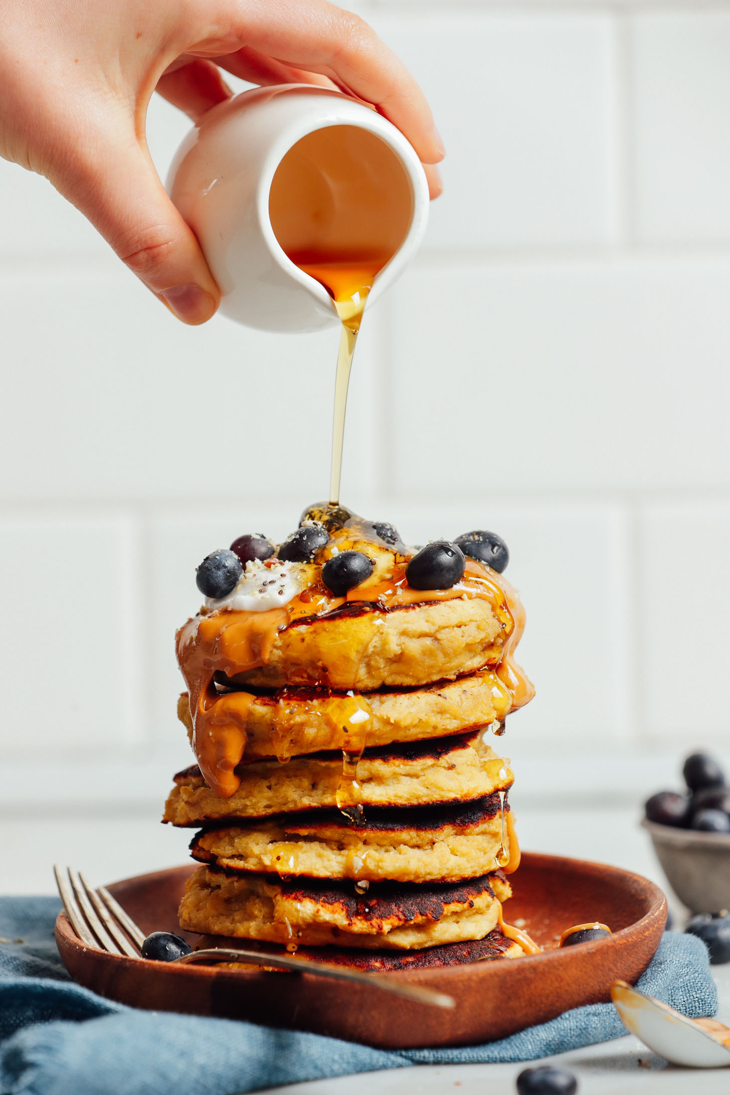 Pouring syrup onto a stack of grain-free dairy-free Banana Pancakes topped with fresh blueberries