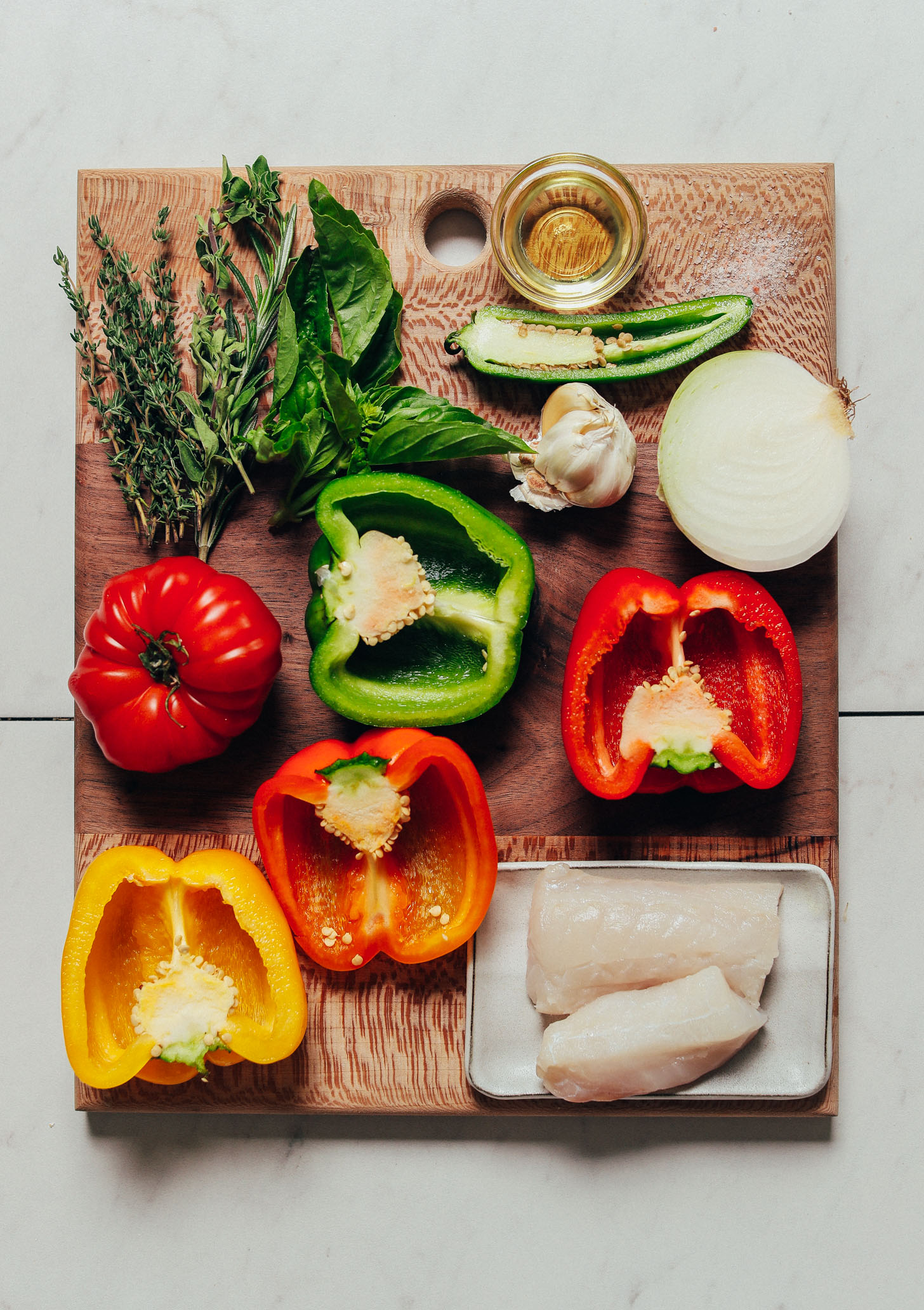 Wood cutting board with bell peppers, onion, fish, herbs, garlic, and other ingredients for making healthy Baked White Fish