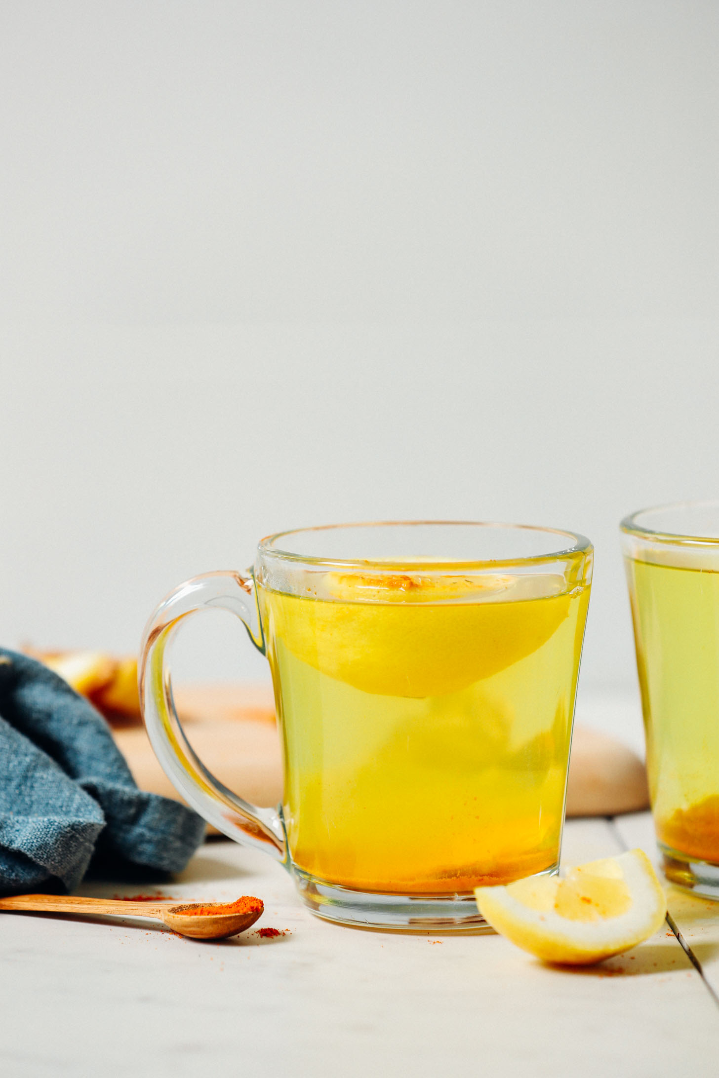straight on image of two mugs of lemon water with ginger, a blue towel on the left, and a lemon wedge in front
