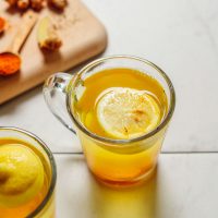 Two glasses of our detoxifying Ginger Lemon Water recipe beside fresh ginger and ground turmeric used to make them