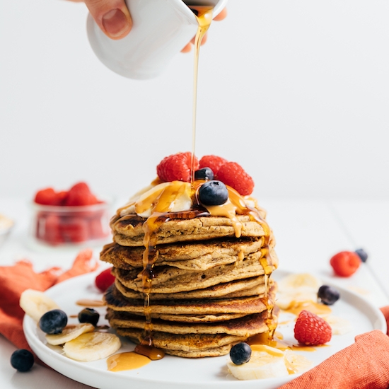 Drizzling maple syrup onto a stack of gluten-free vegan Peanut Butter Pancakes topped with fresh fruit