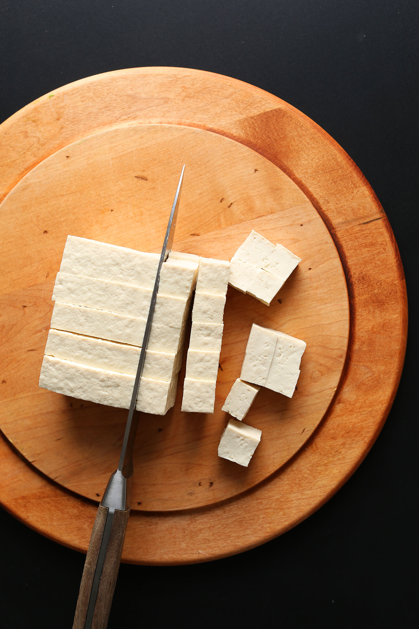 Overhead image of firm tofu on a cutting board being cubed with a large knife