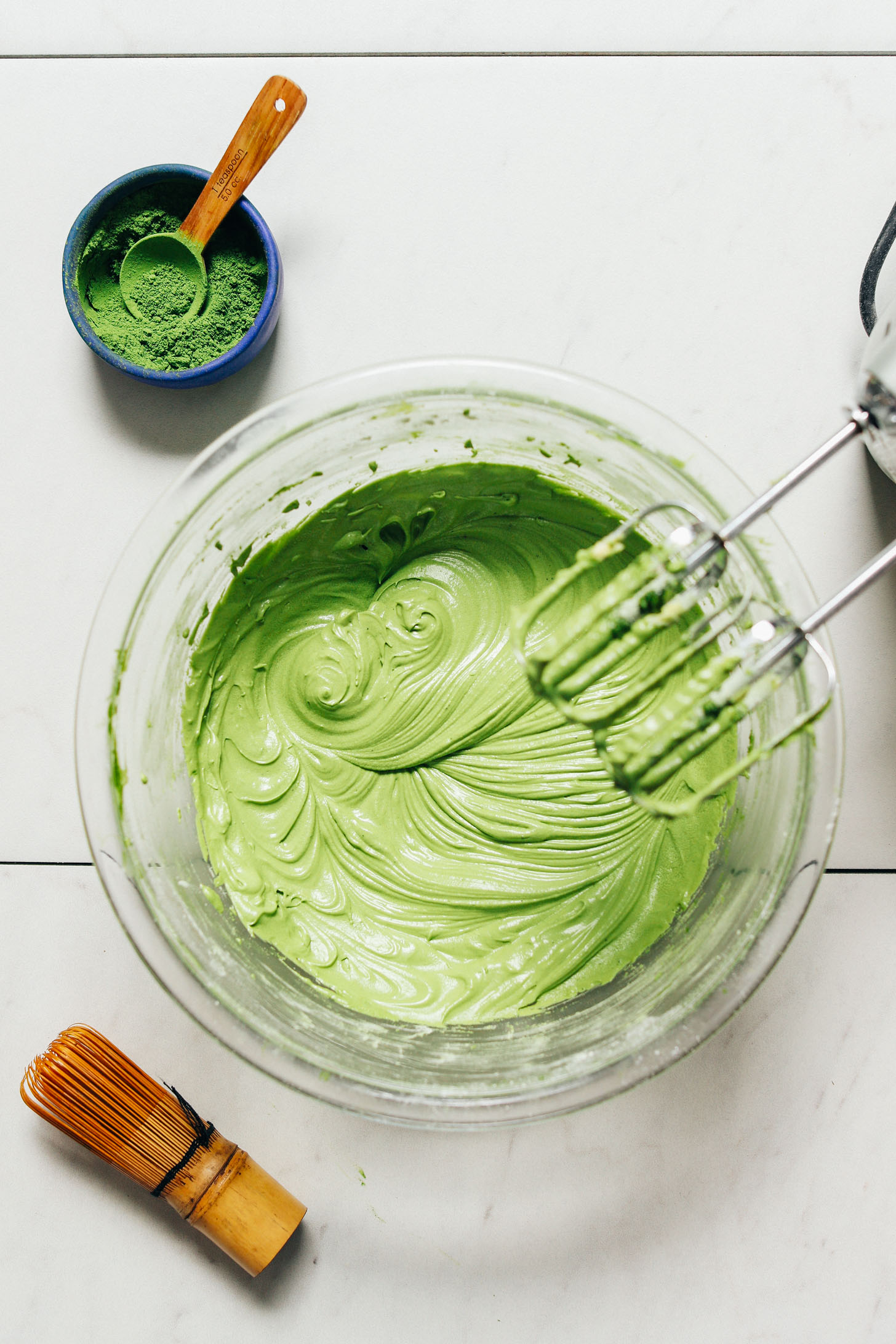 Bowl of matcha buttercream frosting beside a tin of culinary matcha, a bamboo whisk, and a hand mixer