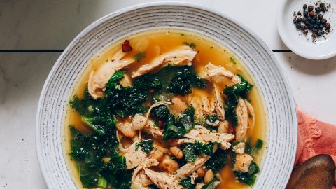 Bowl of 1-Pot Chicken Soup with White Beans and Kale beside ingredients used to make it