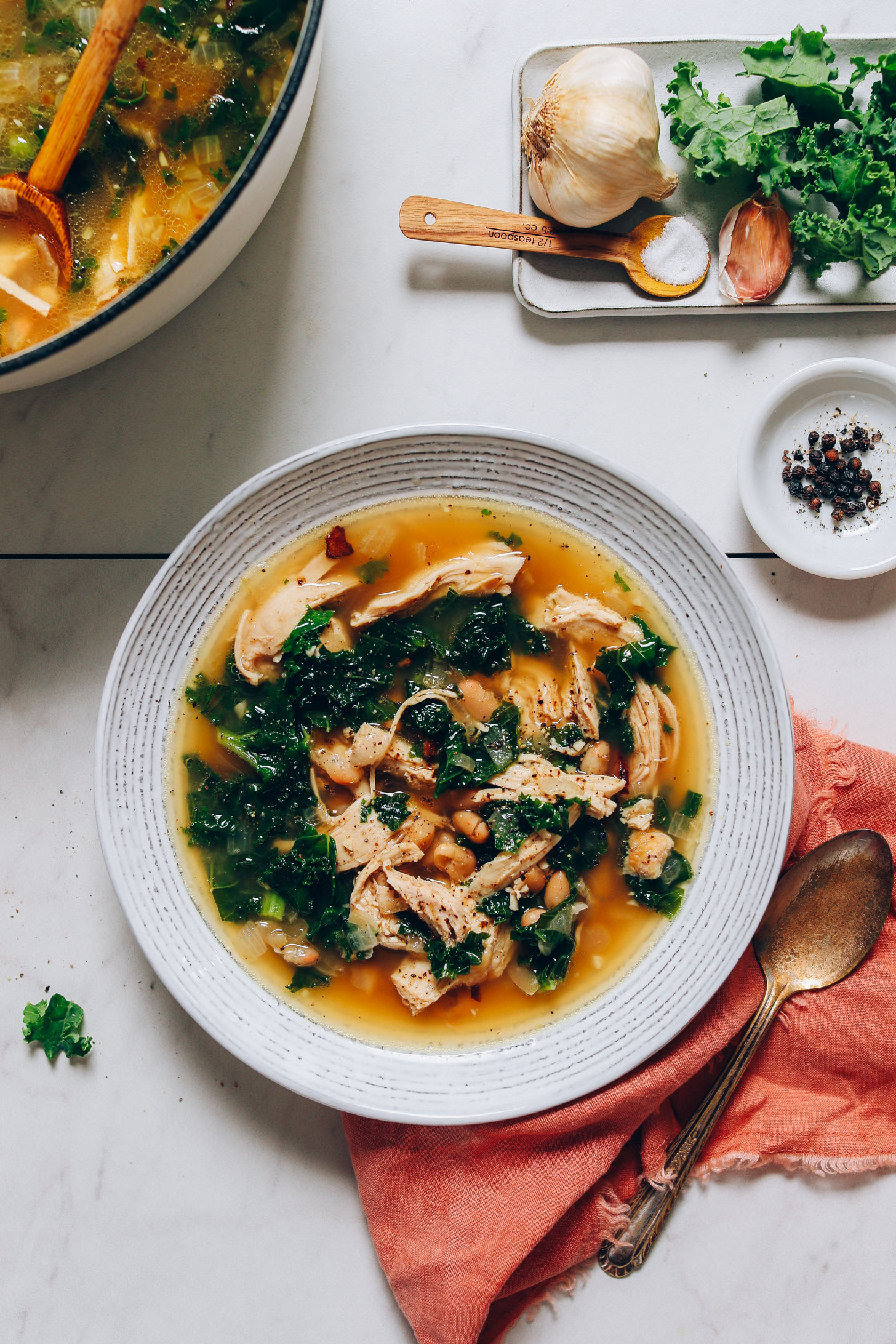 Bowl of 1-Pot Chicken Soup with White Beans and Kale beside ingredients used to make it