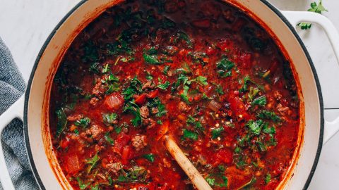 Wooden spoon resting in a pot of Tuscan Beef Soup next to dairy-free parmesan and parsley for serving