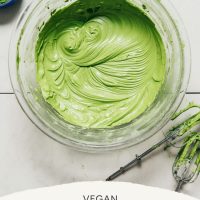Bowl of Vegan Matcha Buttercream Frosting beside a wooden spoon of matcha and mixer wands