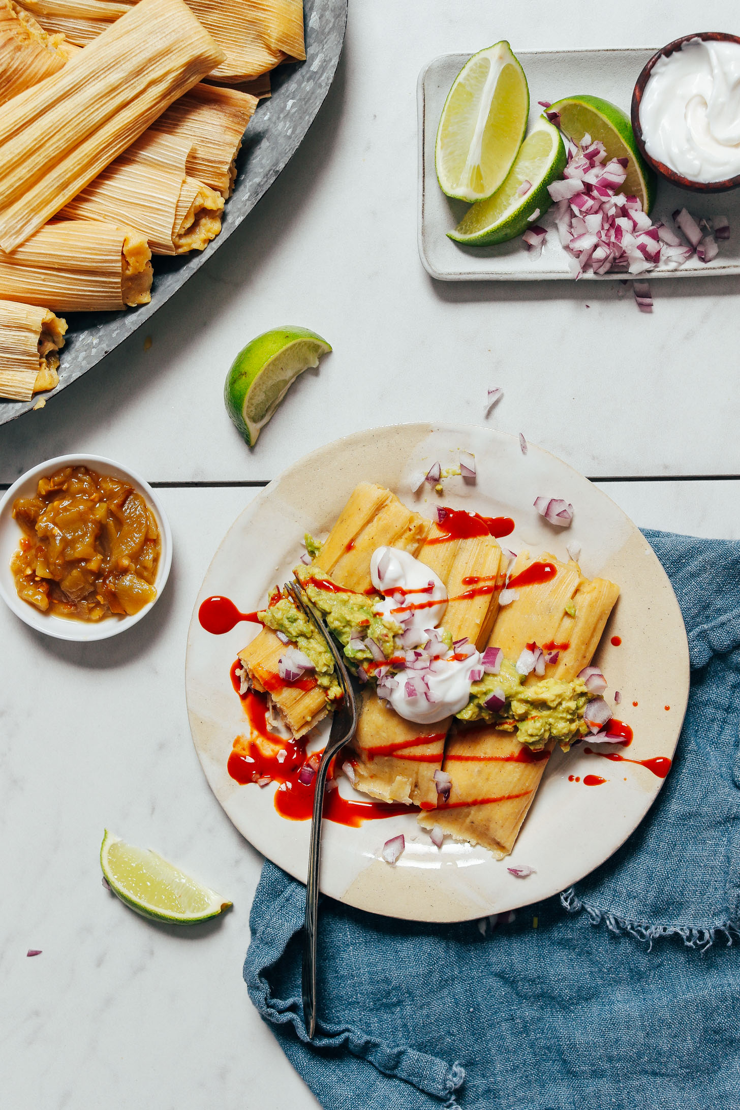 Fork cutting into tamales topped with guacamole, hot sauce, red onion, and sour cream