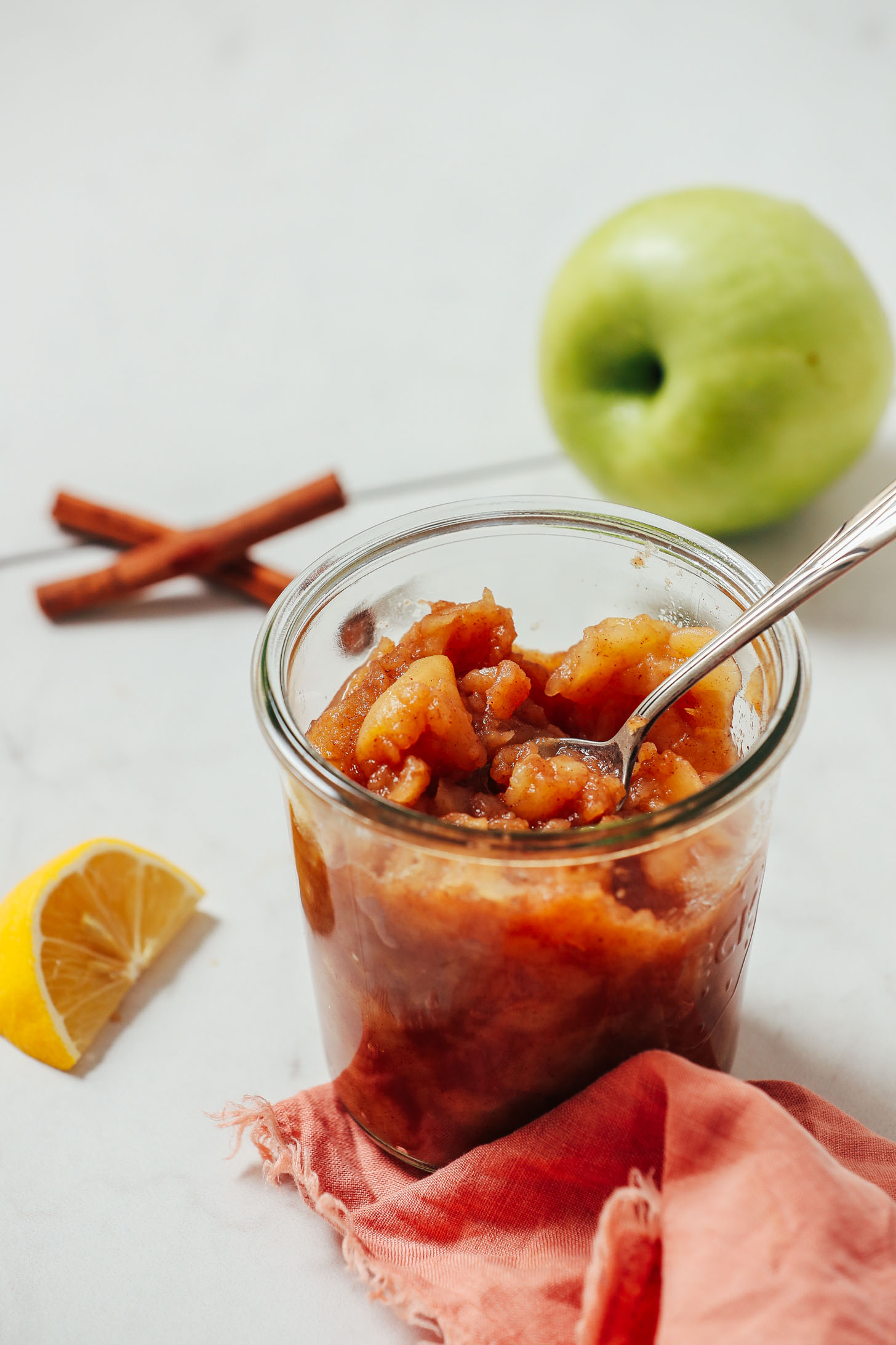 Spoon in a jar of homemade applesauce beside a lemon wedge, cinnamon sticks, and an apple