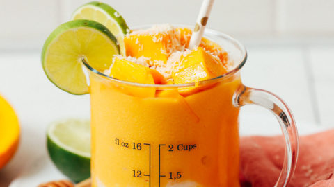 Measuring glass of our mango lassi smoothie topped with frozen mango chunks and sliced lime