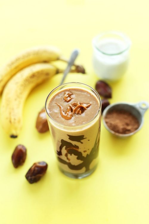Glass of our Peanut Butter Banana Chocolate Shake next to dates, cocoa powder, banana, almond milk, and nut butter