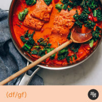 Wooden spoon resting in a pan of red curry with salmon and kale