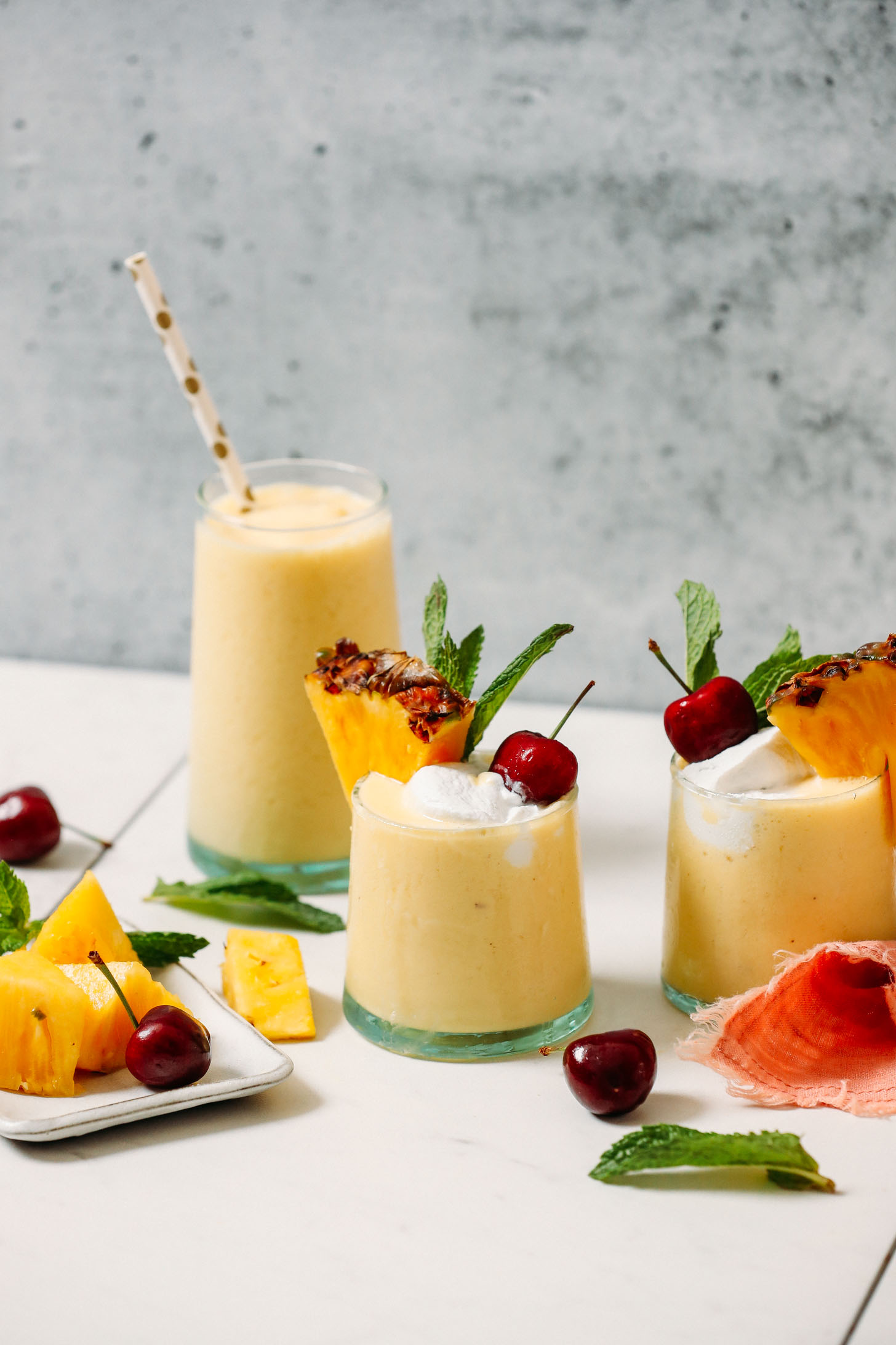Glasses of our piña colada smoothie recipe surrounded by mint, fresh pineapple, and cherries