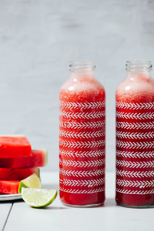 Two bottles of watermelon juice next to freshly sliced watermelon and a lime wedge