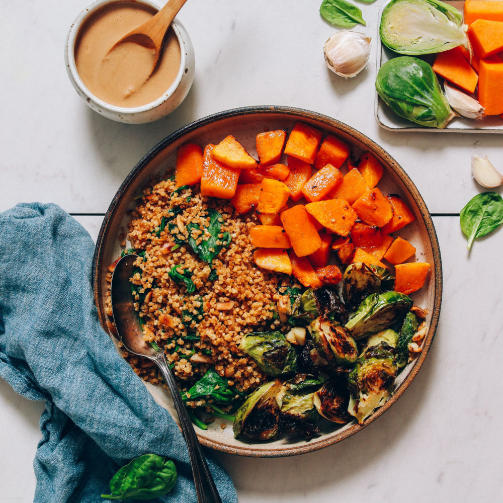 Bowl of roasted butternut squash, miso brussels sprouts, and millet next to tahini sauce, garlic, and vegetables