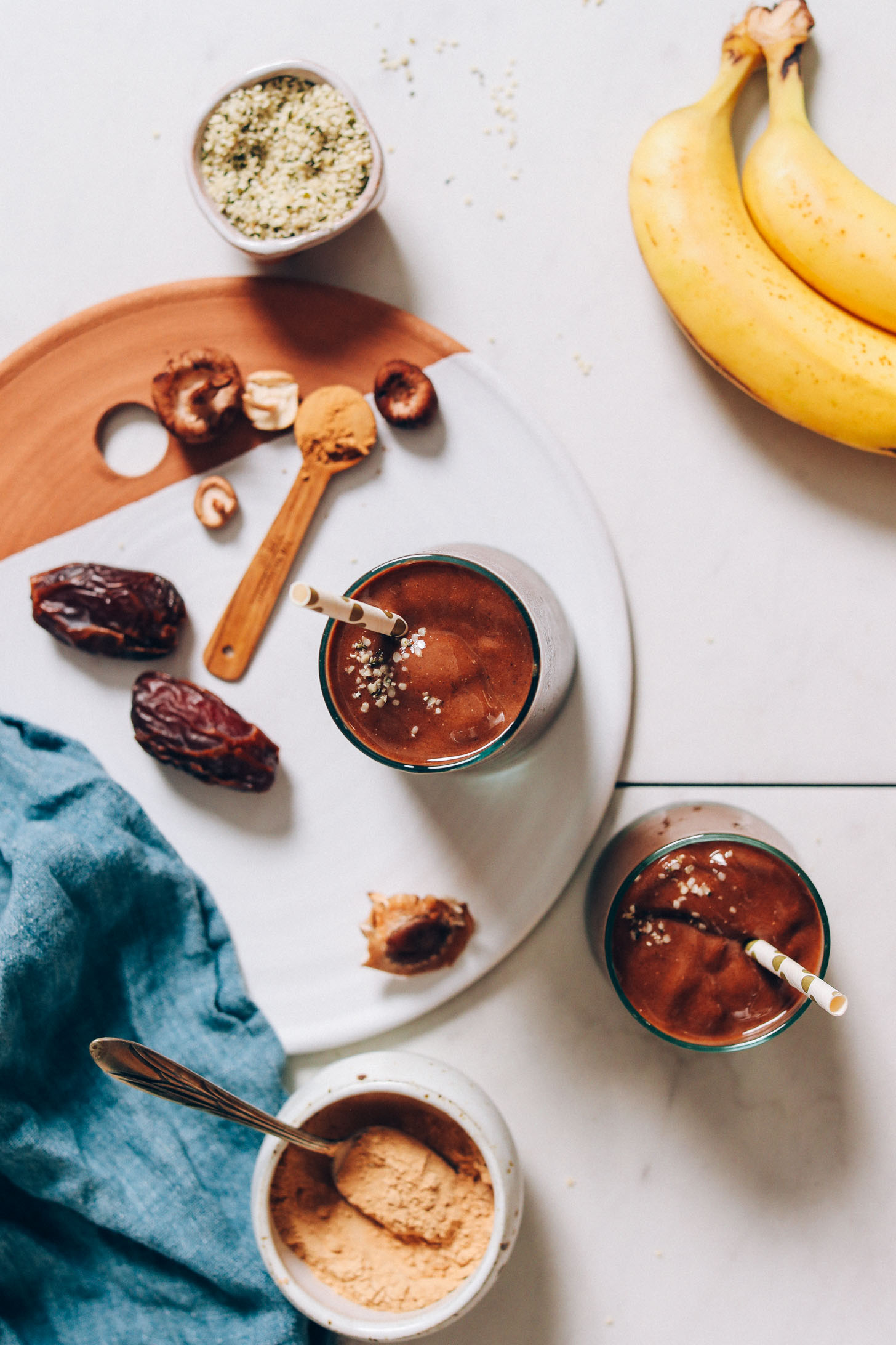 Uma dose de cima para baixo de dois copos do nosso Batido de Chocolate e Banana Adaptogênico ao lado dos ingredientes usados ​​para fazê-lo