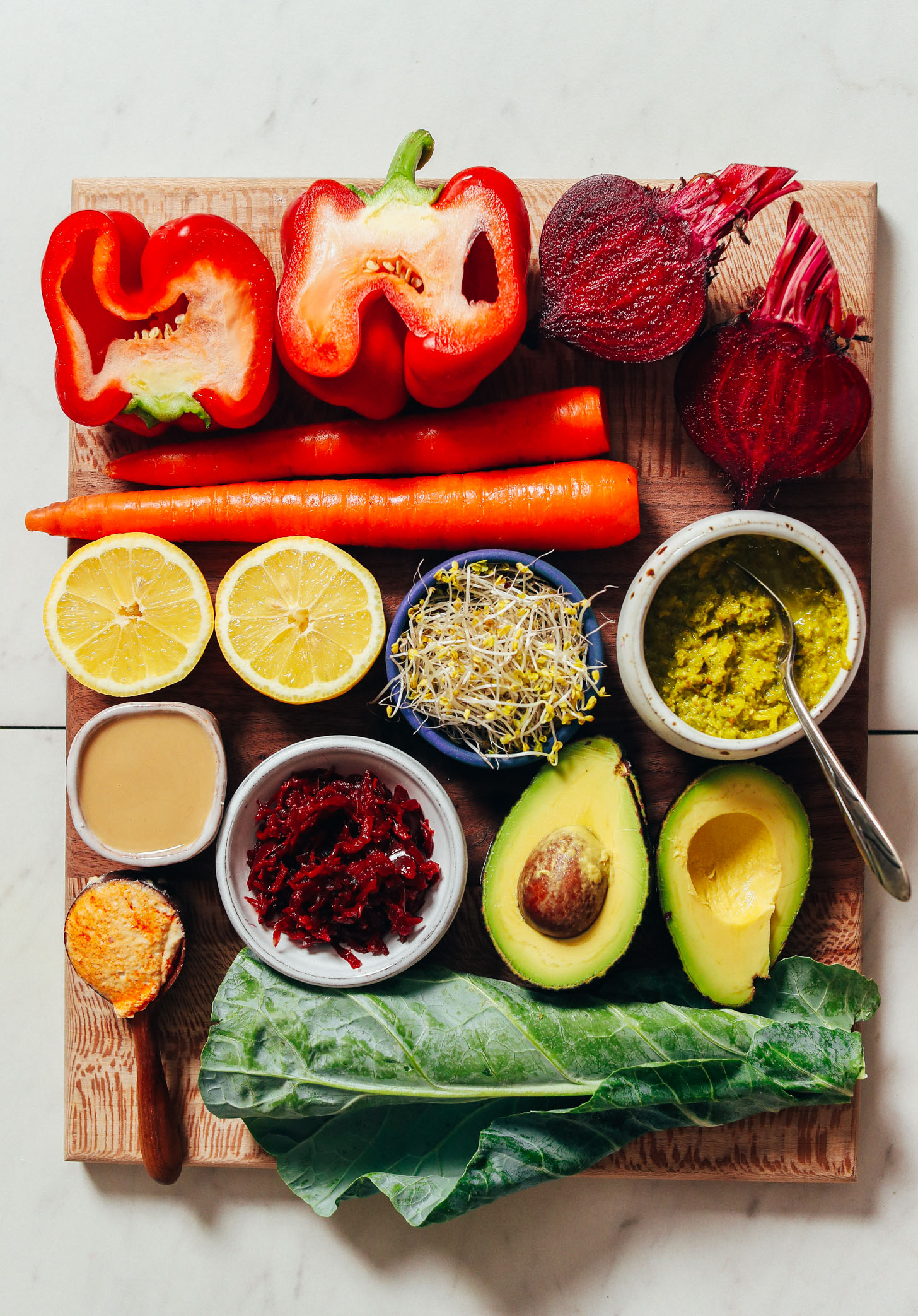 Wood cutting board with bell pepper, carrot, beet, lemon, sprouts, avocado, hummus, tahini, green curry paste, and collard greens