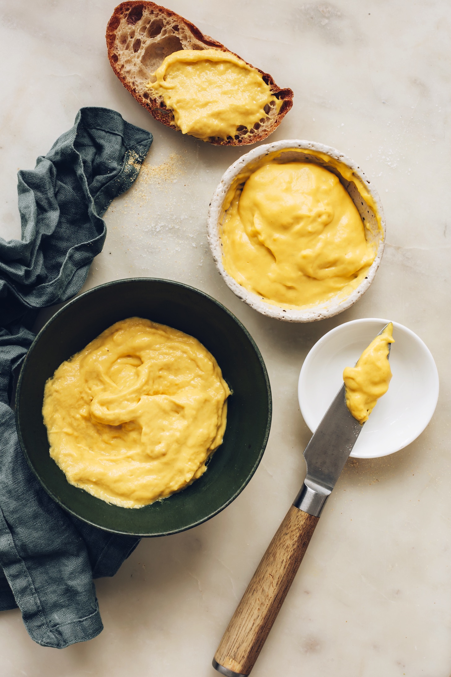 Slice of sourdough bread next to bowls of our easy vegan cheddar cheese recipe