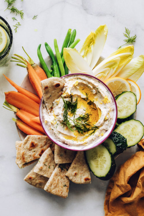 Bowl of white bean hummus surrounded by pita bread and fresh vegetables for dipping