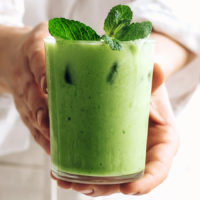Hands holding a glass of our iced matcha latte topped with fresh mint leaves