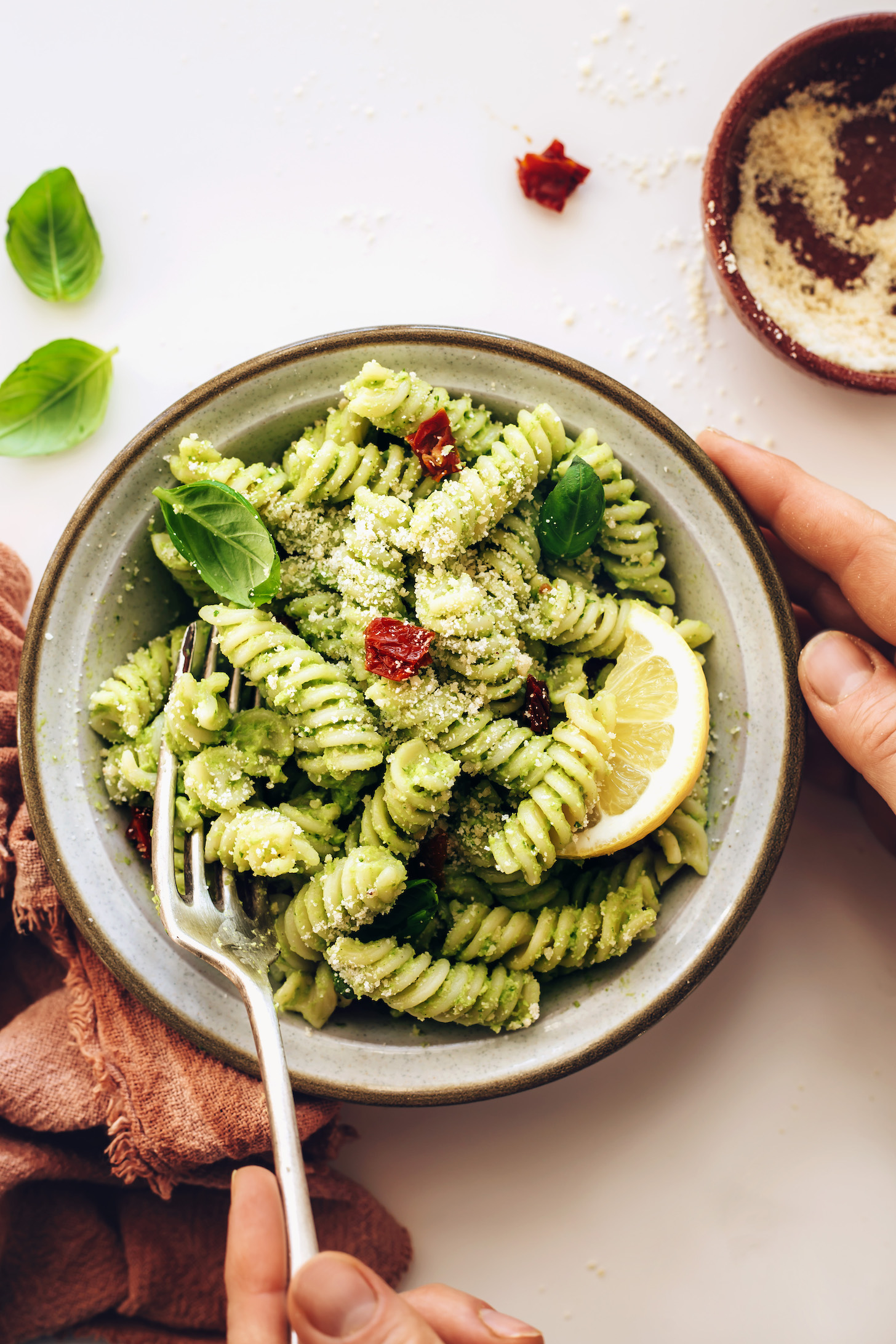 Fork in a bowl of avocado pesto pasta topped with vegan parmesan and sun-dried tomatoes