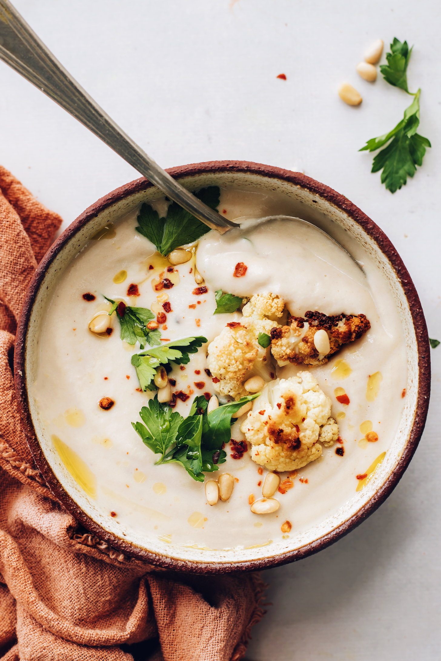 Bowl of creamy roasted cauliflower soup topped with more cauliflower, parsley, pine nuts, and pepper flakes