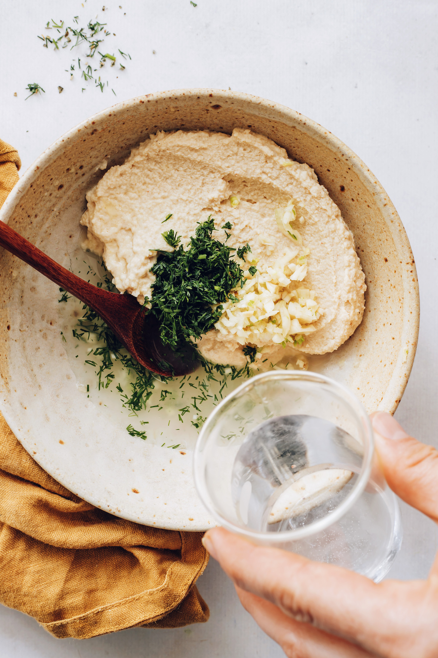 Pouring water into a bowl of hummus, garlic, lemon juice, and dill