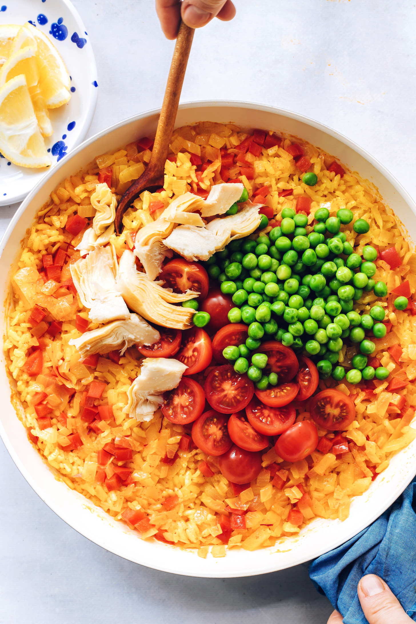 Stirring cherry tomatoes, artichoke hearts, and green peas into a skillet of vegan paella