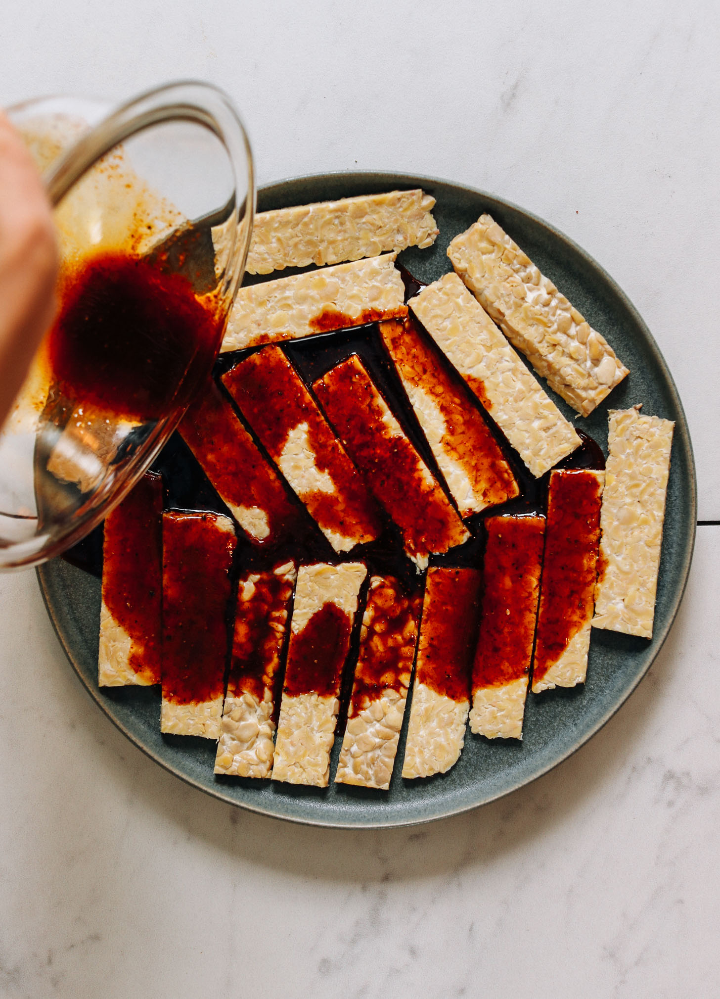 Pouring a tamari-based marinade over strips of thinly sliced tempeh