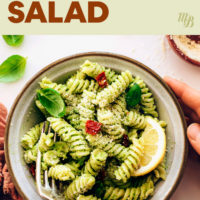 Hands holding a bowl of vegan & gluten-free avocado pesto pasta salad with fresh basil, sun dried tomatoes, and lemon