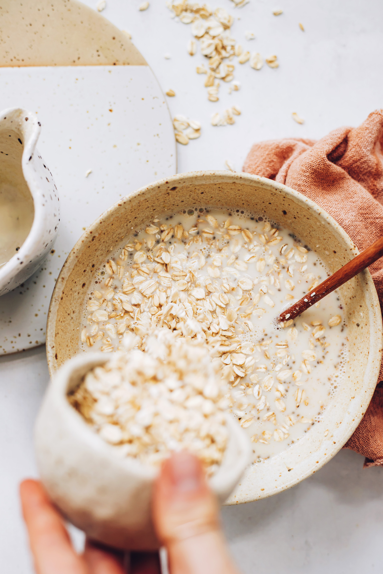 Pouring rolled oats into a bowl of almond milk, chia seeds, and other overnight oat ingredients