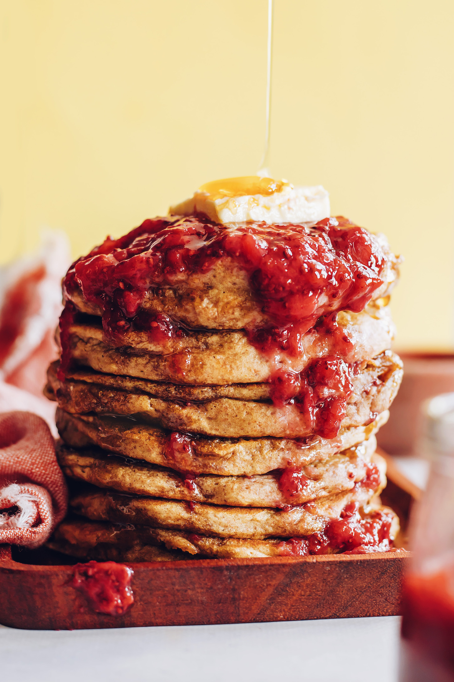 Drizzling maple syrup over a stack of whole grain pancakes with berry compote and vegan butter