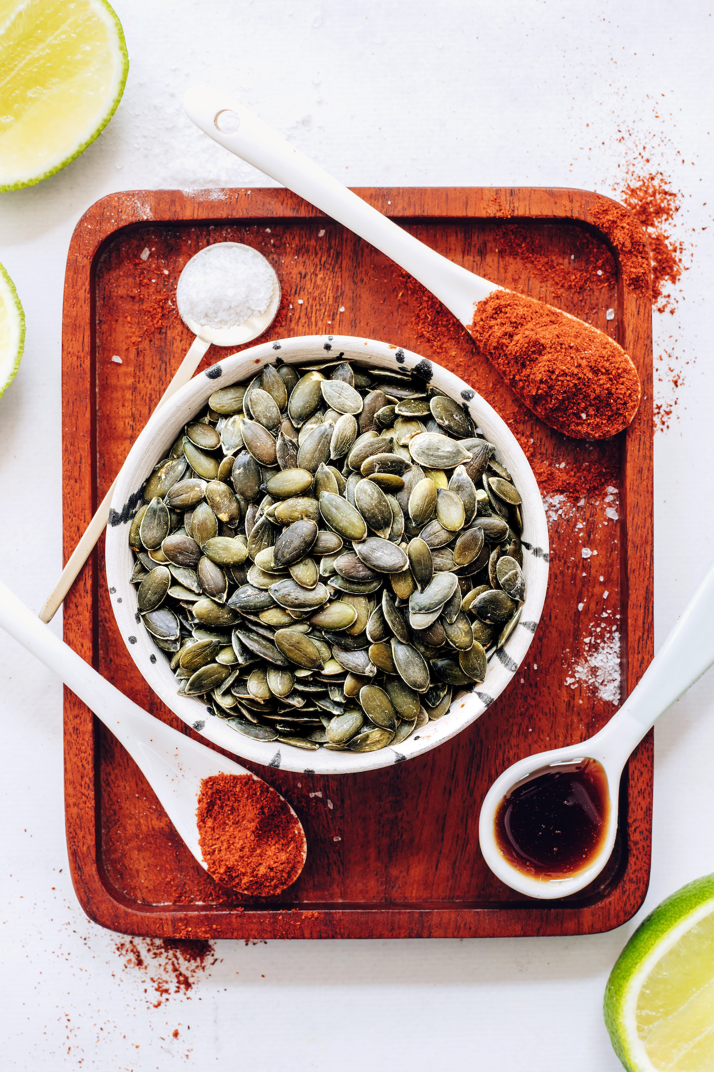 Bowl of raw pumpkin seeds surrounded by spices and limes