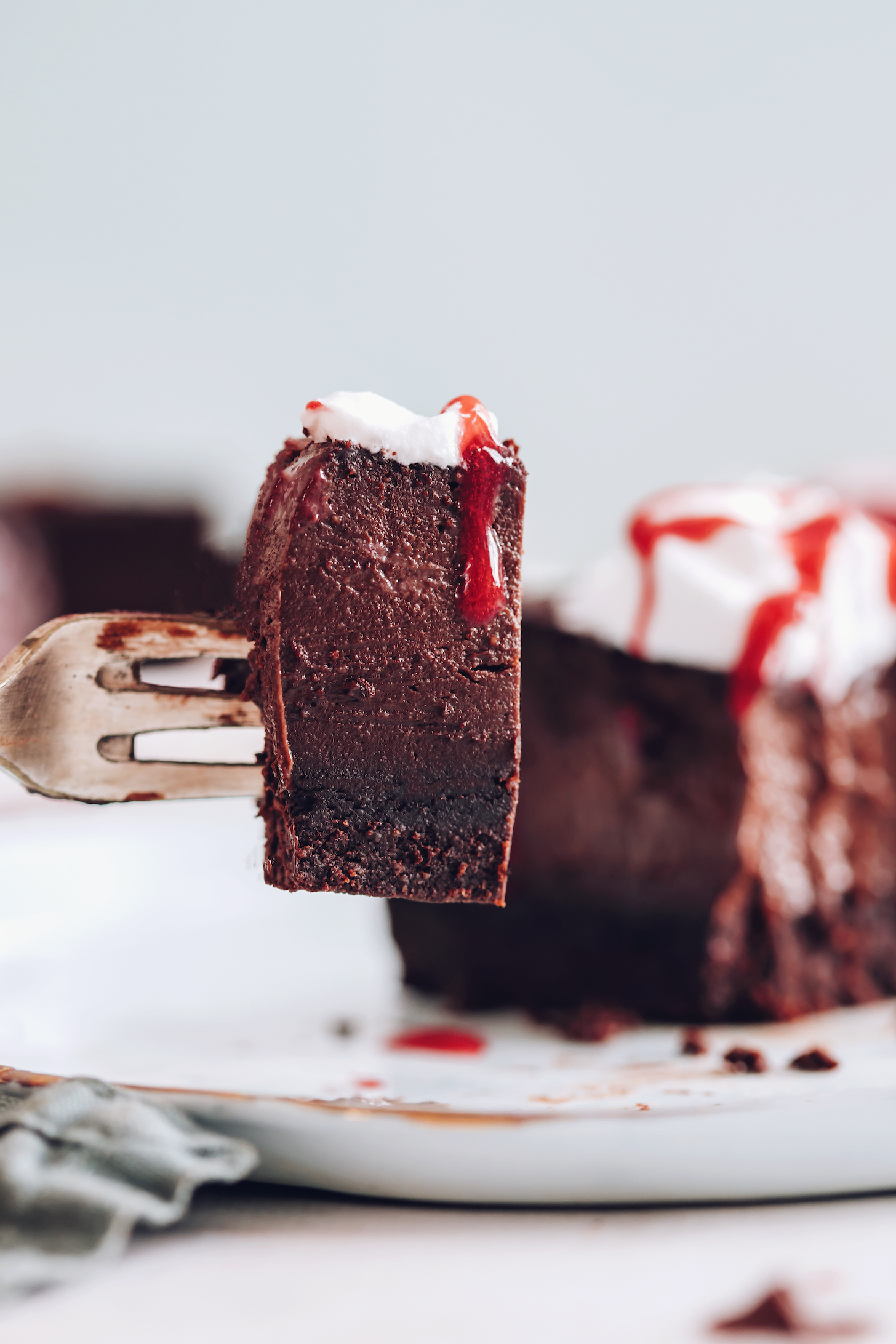 Fork holding up a slice of vegan chocolate cheesecake with coconut whipped cream and raspberry sauce