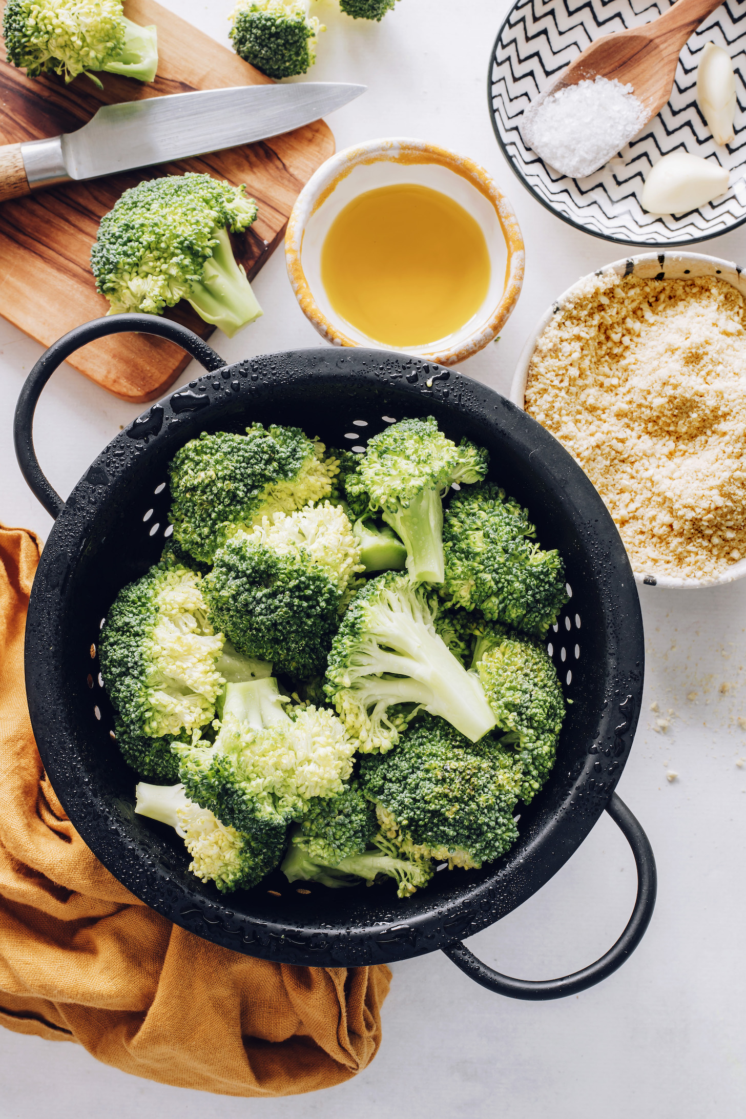 Colander of broccoli florets next to olive oil, vegan parmesan cheese, salt, and garlic