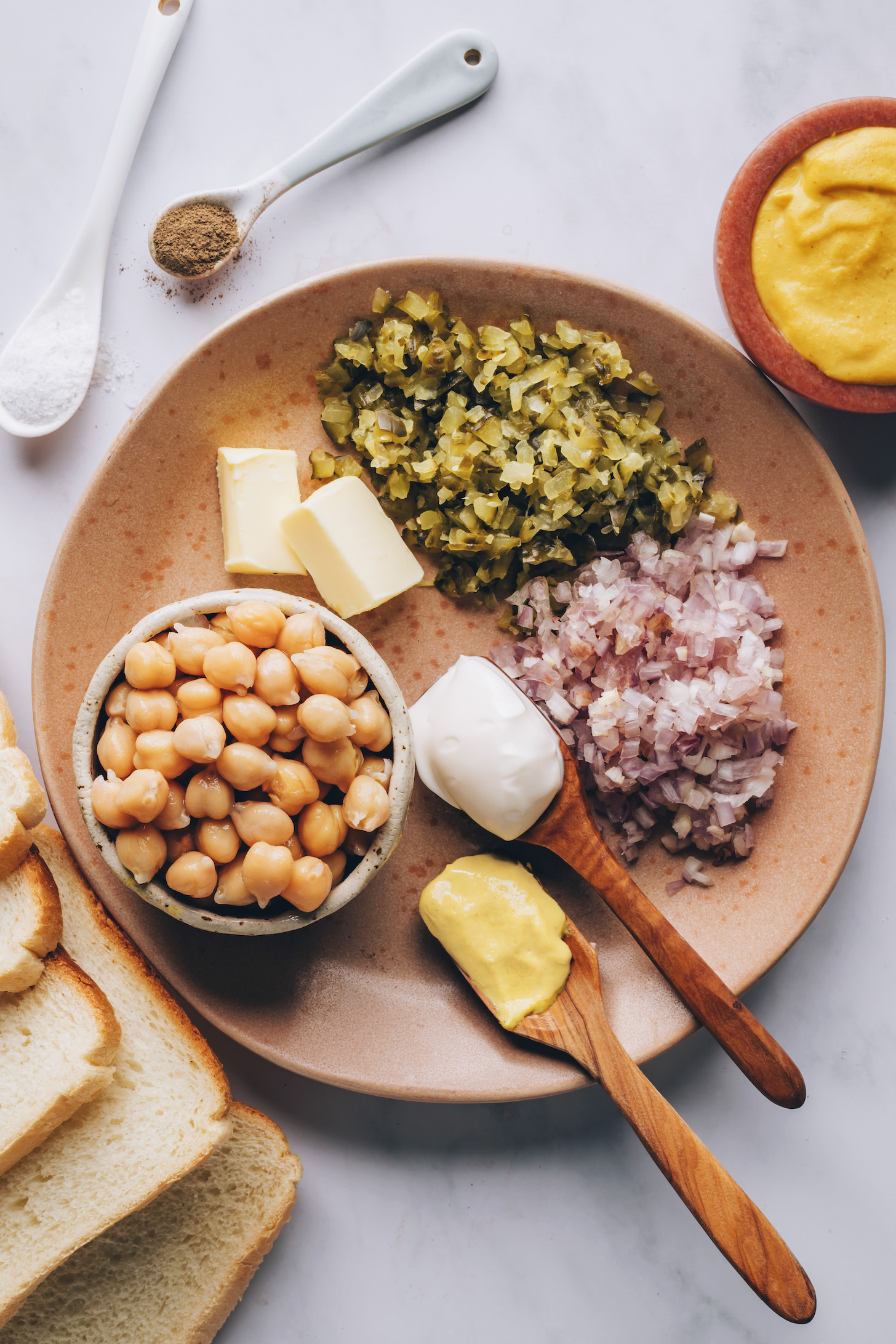 Slices of bread and vegan cheese next to spoons of salt and pepper and a plate with chickpeas, vegan butter, dill pickles, shallot, vegan mayo, and mustard