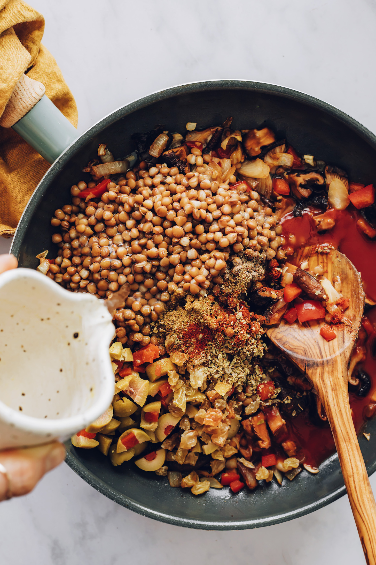 Pouring water over lentils, olives, and spices in a skillet