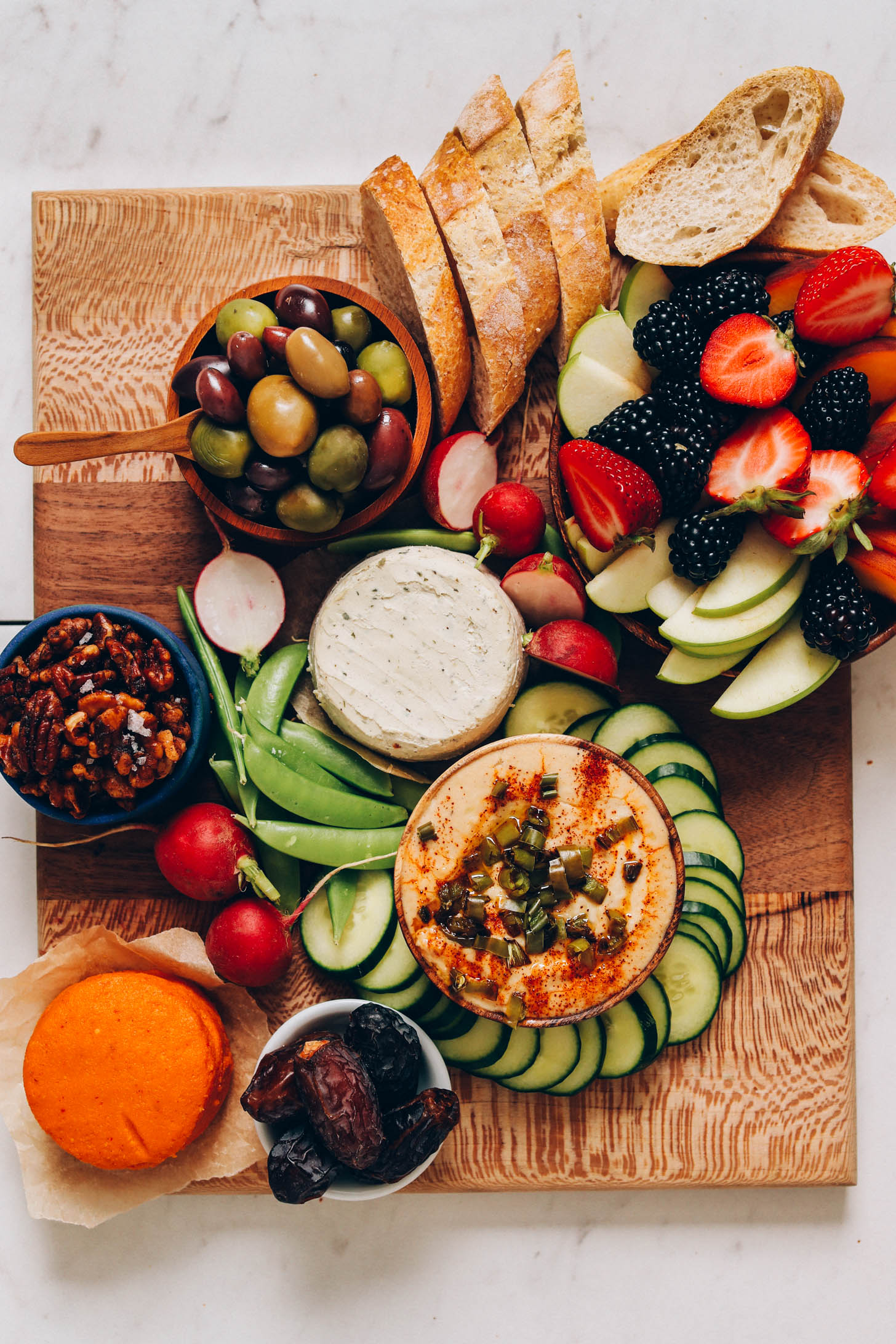 Fresh fruits and veggies assembled on a cutting board around a dip, vegan cheese, and olives